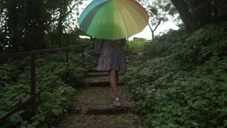Woman-with-rainbow-umbrella-walking-up-a-natural-green-staircase-path,-nature-environment