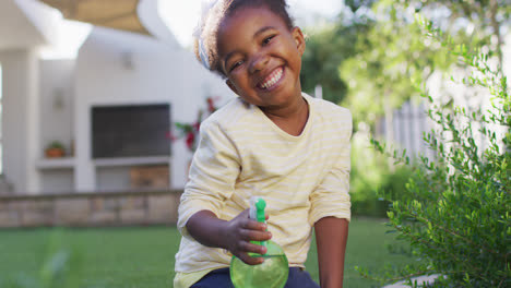 Portrait-of-happy-african-amercian-girl-watering-plants-holding-spray-bottle-in-garden