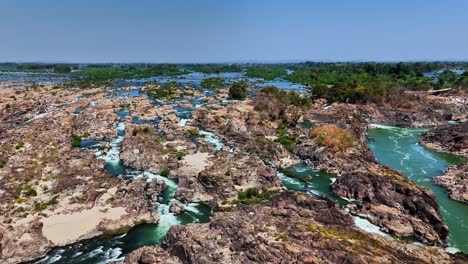 Aerial view over rugged Mekong rapids near Don Det, Laos, showing fractured sandstone and basaltic rock layers split by green waterways, surrounded by tropical vegetation and islets