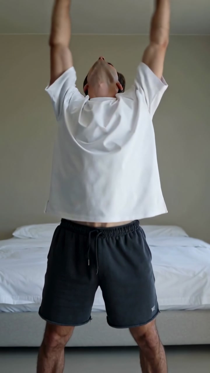 Man starting stretch, turning torso, opening arms, lifting overhead, placing hands on knees by bed