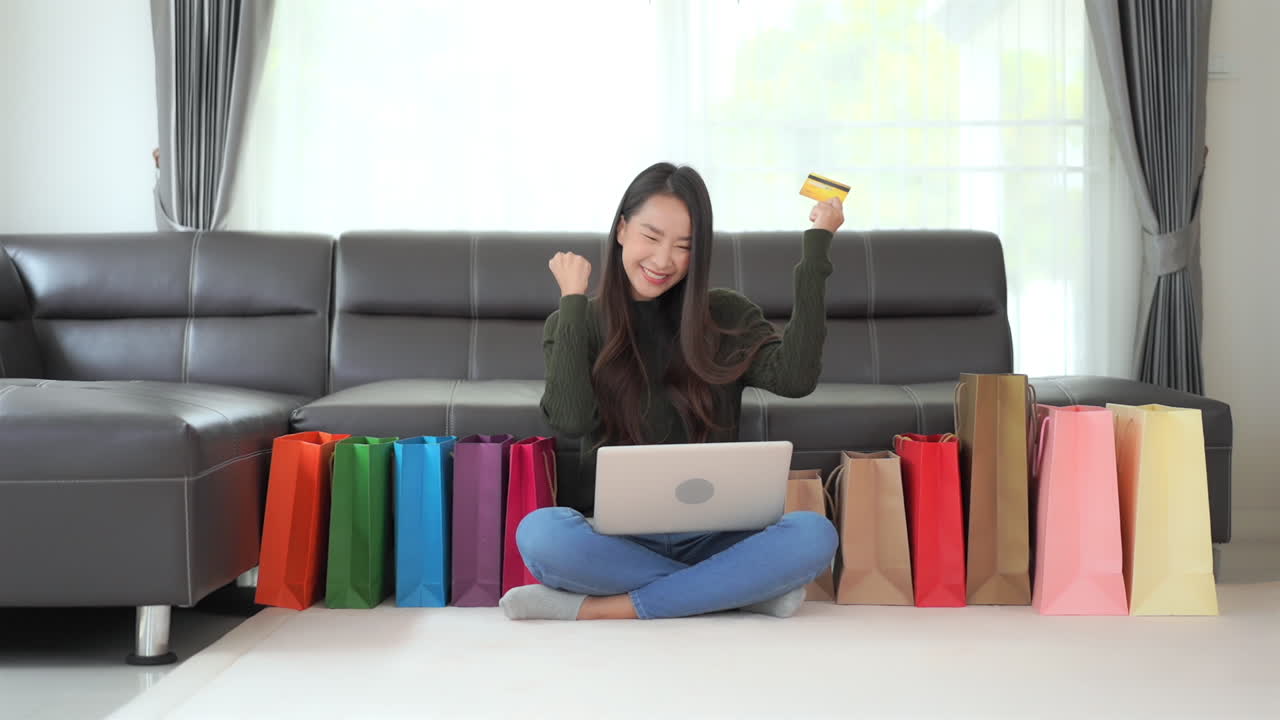 Woman with laptop and credit card smiles and celebrates sitting on floor surrounded by colorful paper bags. Internet shopping in quarantine concept. Happy lady buying more online items Black Friday
