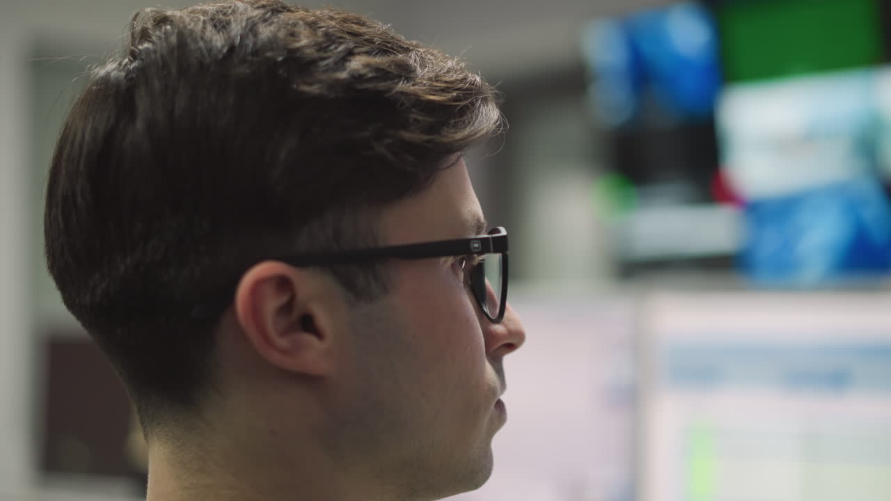 Side profile of male video operator with glasses working in TV control room, focused on tasks, surrounded by screens and high-tech equipment, making technical adjustments