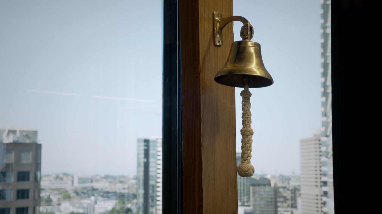 Primo piano di un campanello di servizio in ottone lucido montato su una cornice di legno accanto a una grande finestra, con uno skyline urbano sfocato sullo sfondo