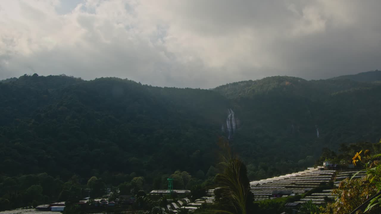 Mountain landscape with waterfall and greenhouses under cloudy sky