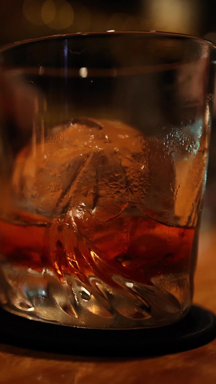 Primo piano estremo di una bevanda ambrata scura con una grande sfera di ghiaccio in un bicchiere.