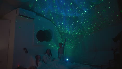 little sister and brother study starry sky using projection on white ceiling. mother and children spend evening lying on cozy bed together. weekend leisure