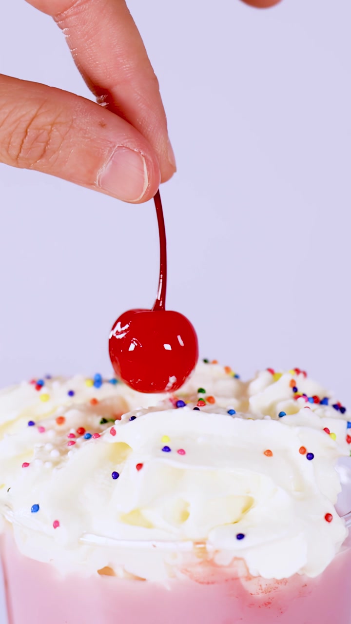 Primo piano verticale di una mano che posiziona una ciliegia rossa su un milkshake con panna montata e codette colorate.