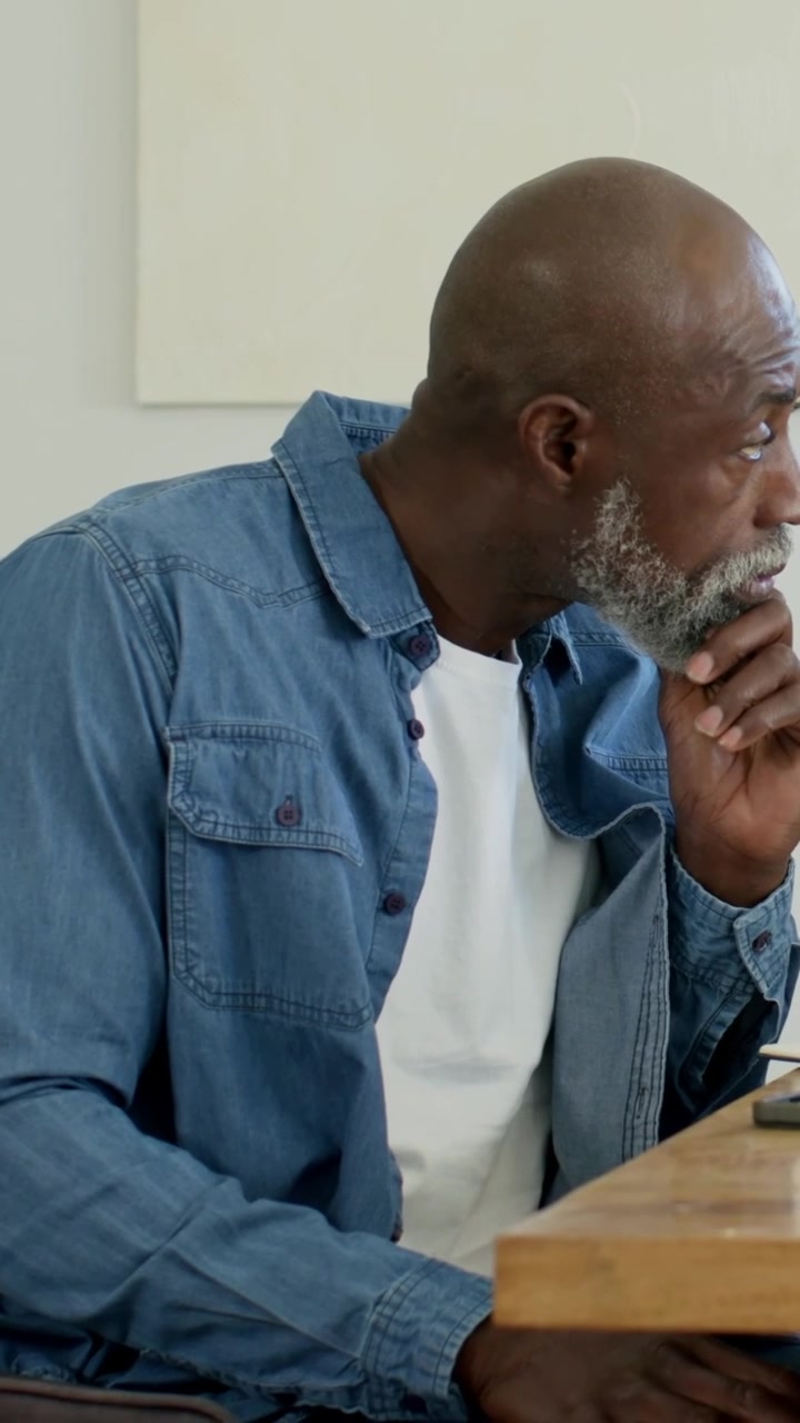 Vertical video: Sitting mature man in denim shirt holding chin and reflecting at desk with phone