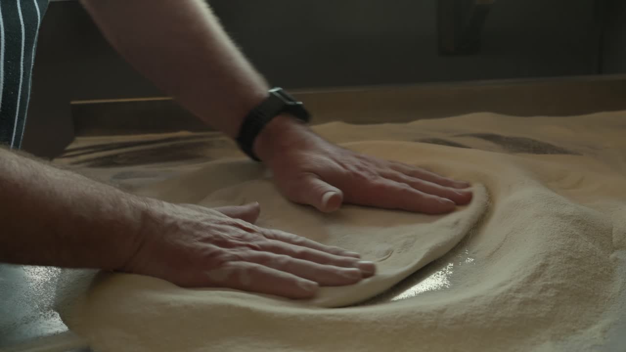 Plano médio-curto de um homem preparando uma massa de pizza com luz suave vinda da janela.