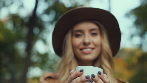 bottom view of caucasian woman in a hat holding a white cup of coffee and looking at the camera in the park in autumn