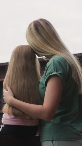 loving mother hugs little daughter sitting on terrace of eco hotel looking at old distant mountains with mist cloud backside view slow motion