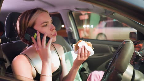 woman in black top eating doughnut while on phone call in parked car at night, looking contemplative, soft lighting, shopping bag visible in backseat, with a slight glow from nearby vehicles