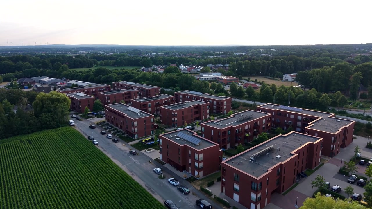New brick apartment complex spreads beside cornfield in American suburb, multiple condo buildings with rooftop solar panels line paved streets near parked cars and trees. Aerial drone flyover shot