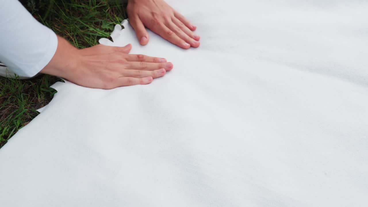 피크닉 준비를 하는 동안 젊은 남자가 부드러운 흰색 천을 초록색 잔디 위에서 조심스럽게 펼치는 모습이에요. 자연광 아래에서 세심한 움직임과 텍스처 디테일을 보여주며 준비하는 느낌을 주네요.