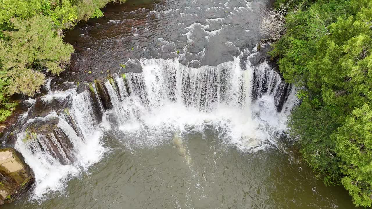 Drone ascends above cascading waterfall, revealing forested riverbanks in bright natural daylight