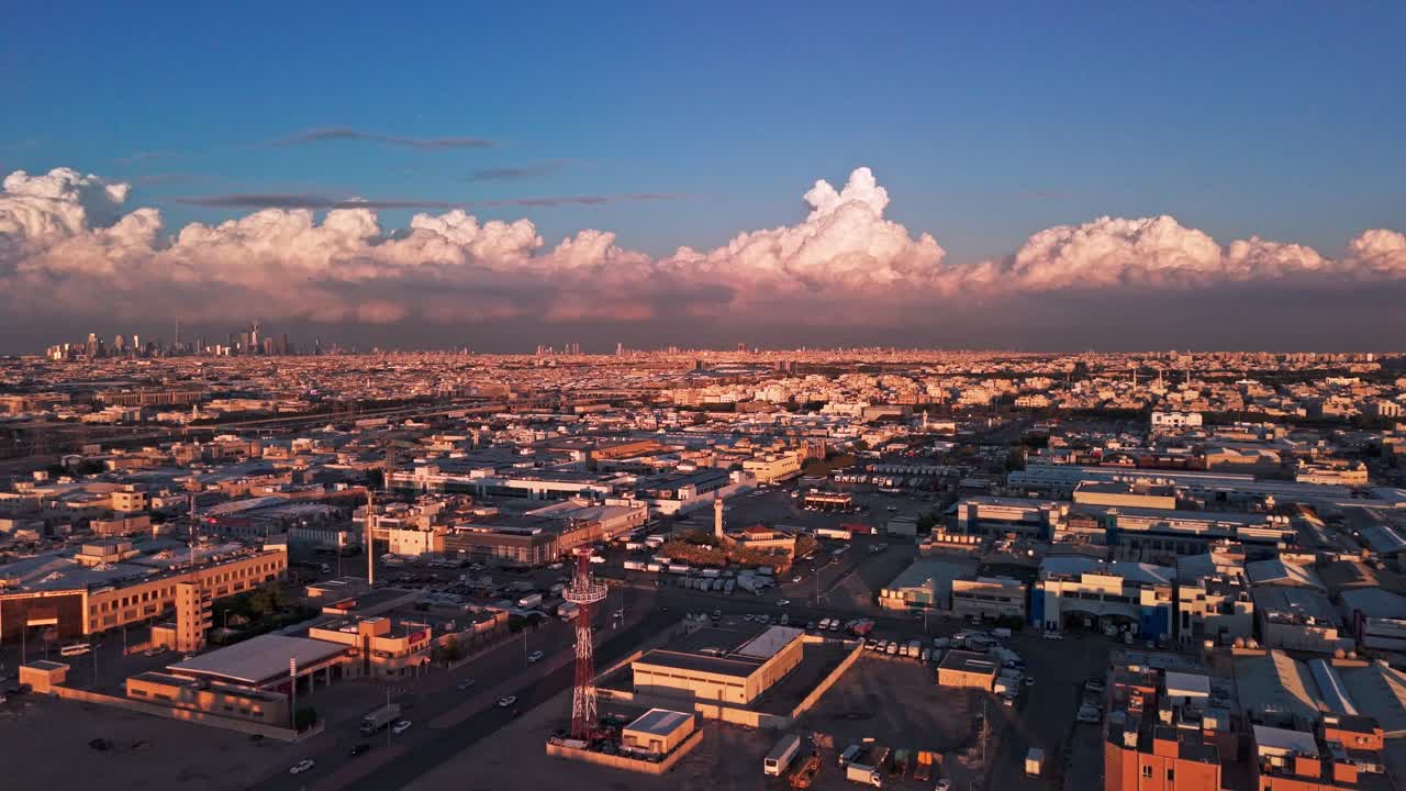 Bredt droneview mod Kuwait City: gyldent timelys, tårnhøje cumulonimbuslinjer, kystlinje, tæt boligområde, motorveje, master og varehuse – rent etablerende billede til rejsebrug