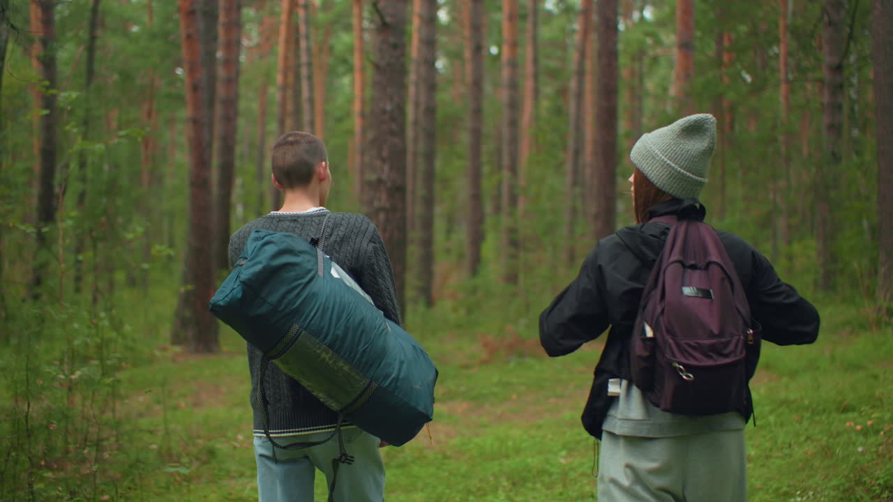 Siblings trekking through forest path surrounded by trees and greenery as girl adjusts backpack strap and scans environment while walking, both wearing casual clothing and carrying camping equipment