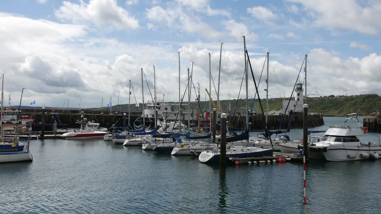 Boats and sailboats moored in the marina at Scarborough, North Yorkshire in England, with a white lighthouse and green hills visible under a partly cloudy sky