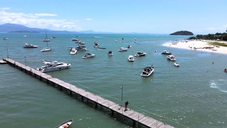 escena aérea de aviones no tripulados de bajo vuelo sobrevolando lanchas rápidas, yates y barcos de lujo con muelle en playa paradisiaca en canajure florianópolis santa catarina