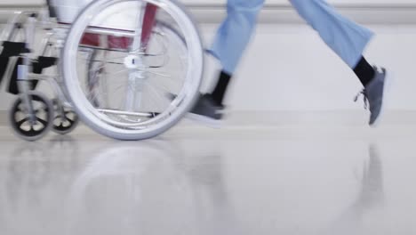 Unterteil-Eines-Afroamerikanischen-Männlichen-Arztes,-Der-Mit-Rollstuhl-Im-Krankenhausflur-Läuft,-Zeitlupe