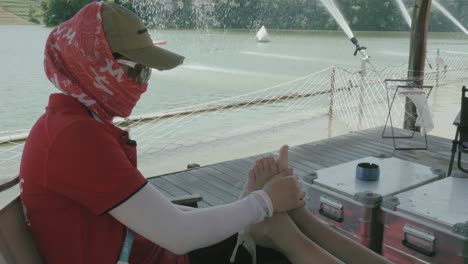 una persona con un sombrero y una cubierta facial está usando hielo para tratar el pie quemado de otro individuo, sentado cerca de una cubierta a la orilla del lago