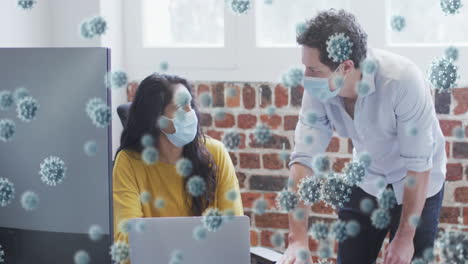 animation of covid 19 cells floating over man and woman wearing face masks, working laptop in office