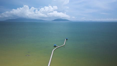 Simple-calm-walking-pier-to-huts-jetting-out-into-the-blue-green-ocean-with-island-on-the-horizon