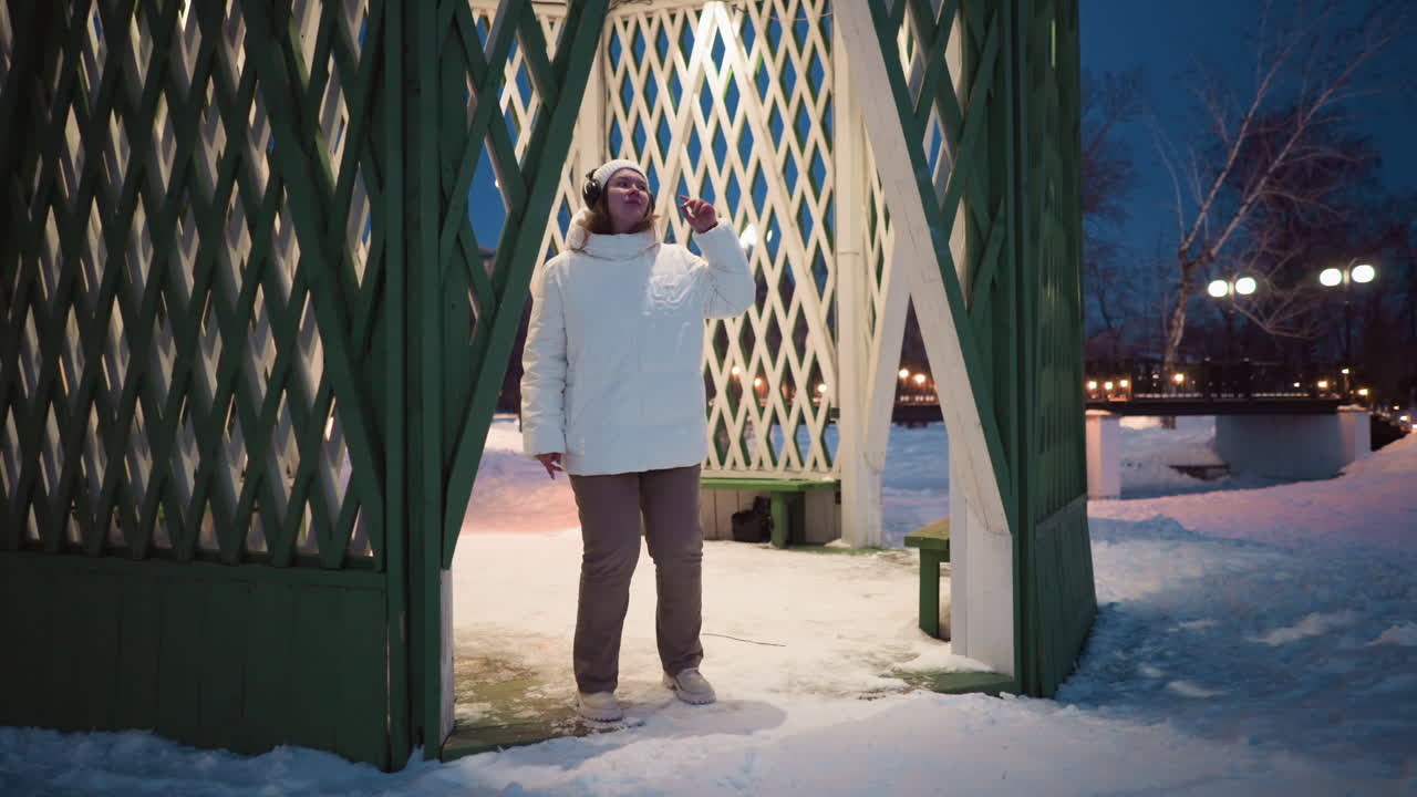 Woman stands at entrance of lighted shelter in snow covered park at night, wearing white puffer coat headphones and beanie, surrounded by decorative lights and bare trees under winter sky