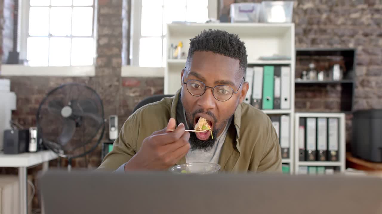 Focused african american casual businessman using laptop and having lunch in office in slow motion
