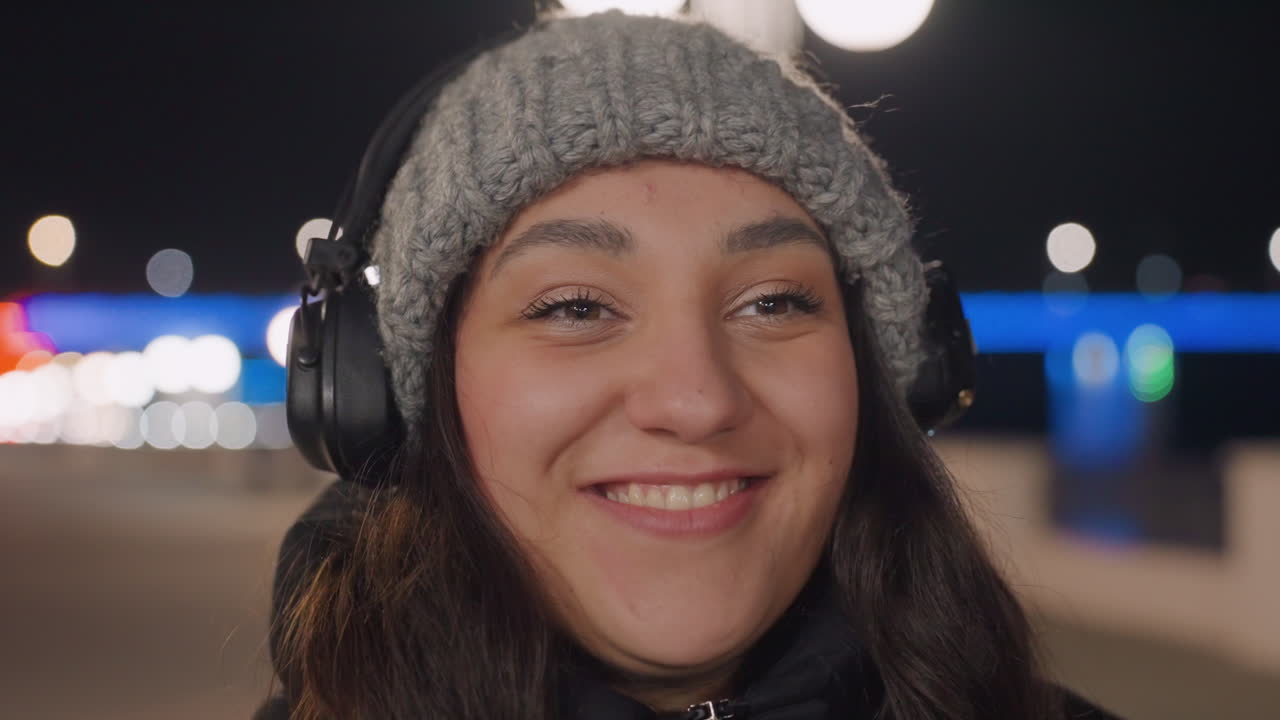 Night street dancer with headphones smiling, knit beanie and sunglasses transition to overear headphones as she grooves under lamp posts and neon bridge lights candid closeup captures rhythm, joy