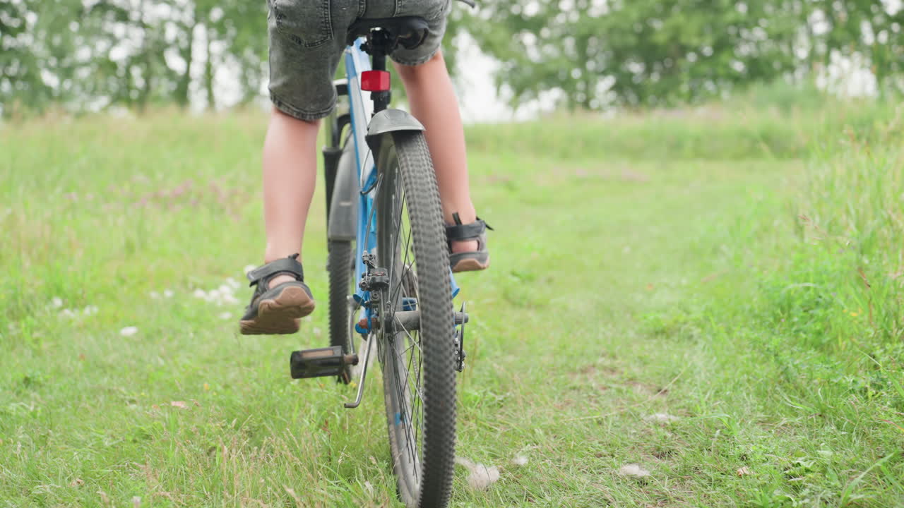잔디밭에서 파란색 자전거를 타는 백인 아이, 다리와 페달의 클로즈업은 망설이는 듯한 추진과 균형 연습을 보여주고, 샌들과 흙 묻은 신발, 보이는 체인과 기어, 여름 목초지 배경