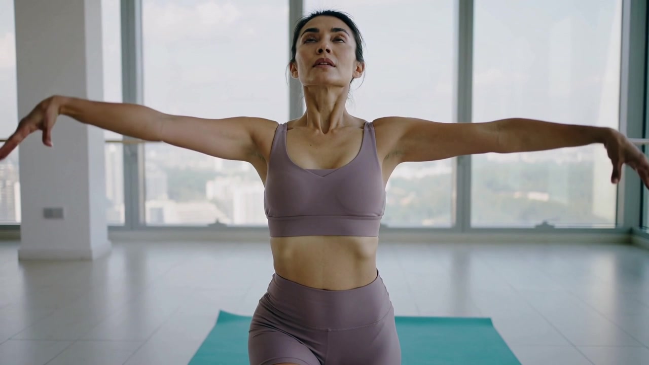Mujer asiática vestida de malva en el centro, levantándose y estirándose hacia el horizonte en un estudio de piso alto sobre una esterilla verde azulado.