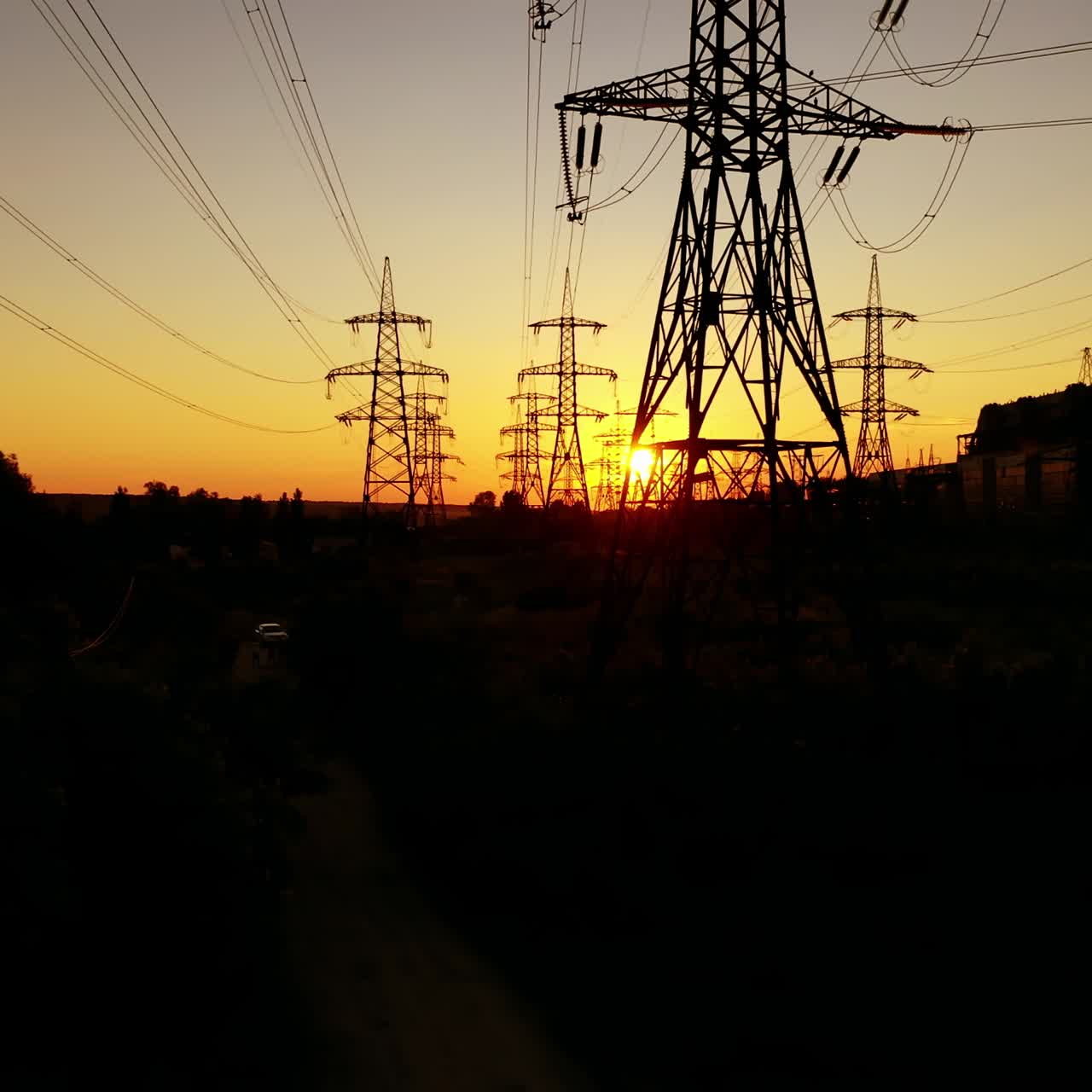 Power station near the river at sunset. Tall silhouettes of high-voltage electric lines in the evening. Electric towers on orange background of the setting sun.