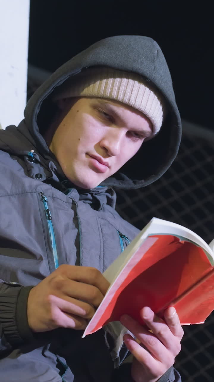 estudiante con capucha y gorra apoyado contra un poste de meta metálico al aire libre por la noche, centrado en leer un libro con una valla de malla de fondo, mirando algo en la distancia en un entorno urbano
