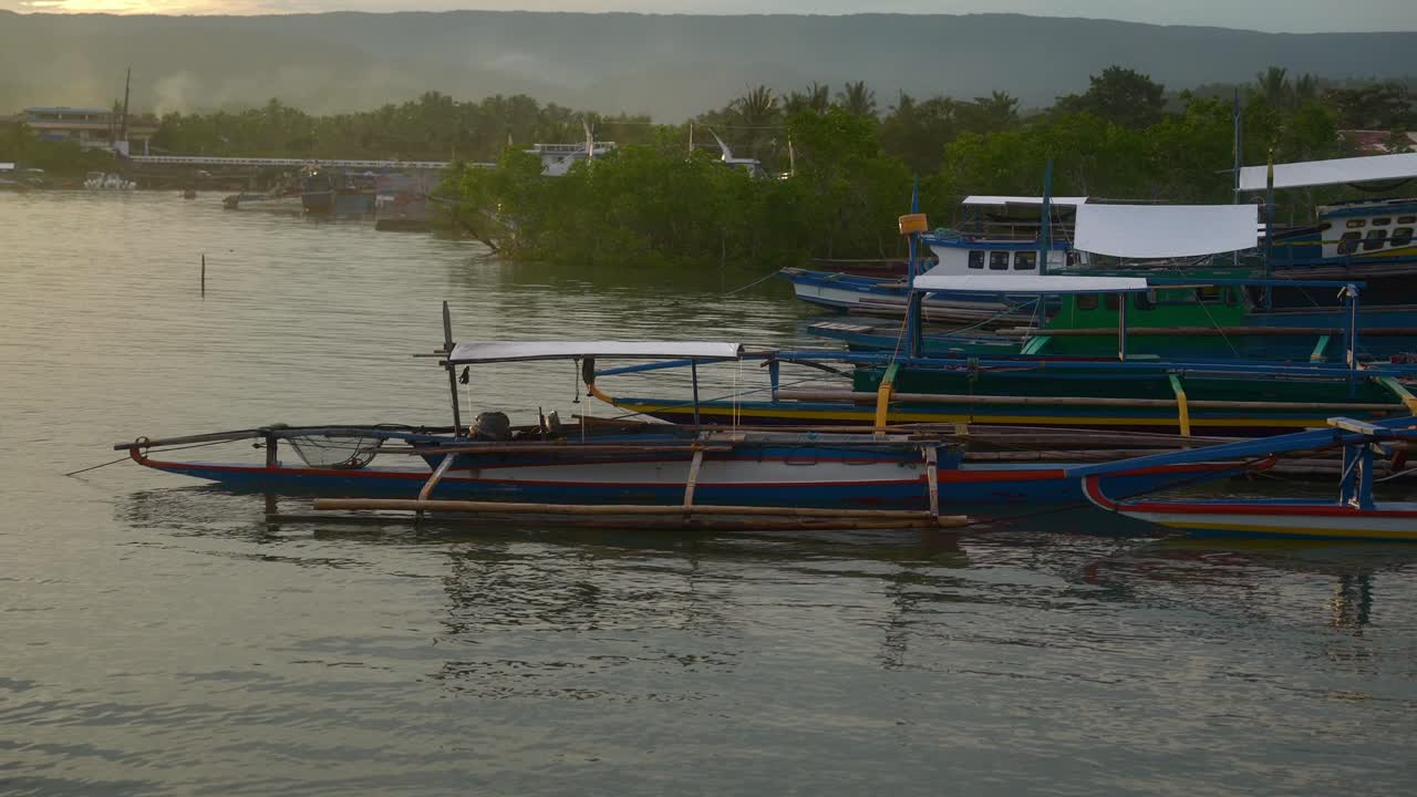A focus shot of a traditional wooden outrigger boat docked with calm waters swaying it at the Mauban Port in Quezon Province Philippines under the golden sunset on the horizon