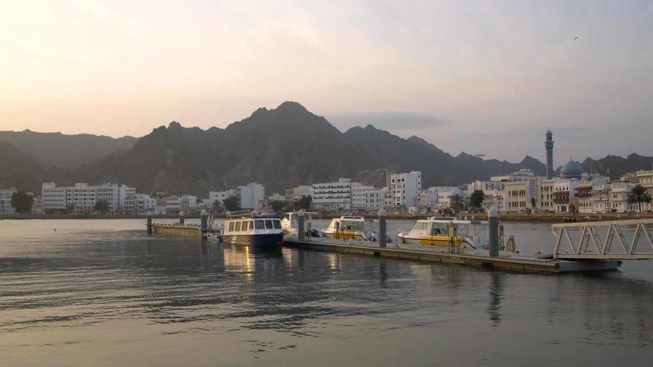 muelle con barcos en mutrah corniche con pájaros volando y montañas en el fondo durante el amanecer