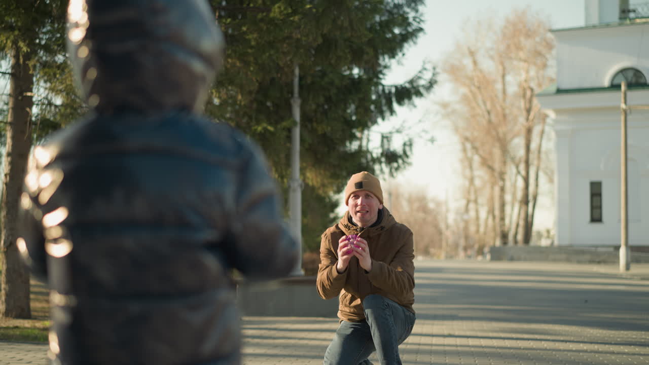 야외에서 놀고 있는 아버지와 아들의 클로즈업, 아이가 핑크색, 반이는 물체를 들고, 배경에는 색 건물이 있고 주변에는 나무가 있습니다.