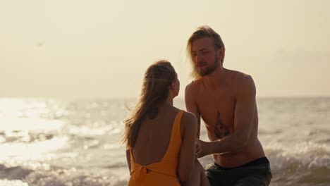a blonde girl in an orange swimsuit kisses her blonde boyfriend with a bare torso in black shorts while sitting on the beach near the sea early in the morning