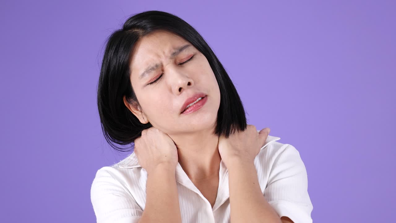 白いシャツを着た若い女性が、ソフトなスタジオ照明の下で首をマッサージし、痛みと疲労を訴えています。