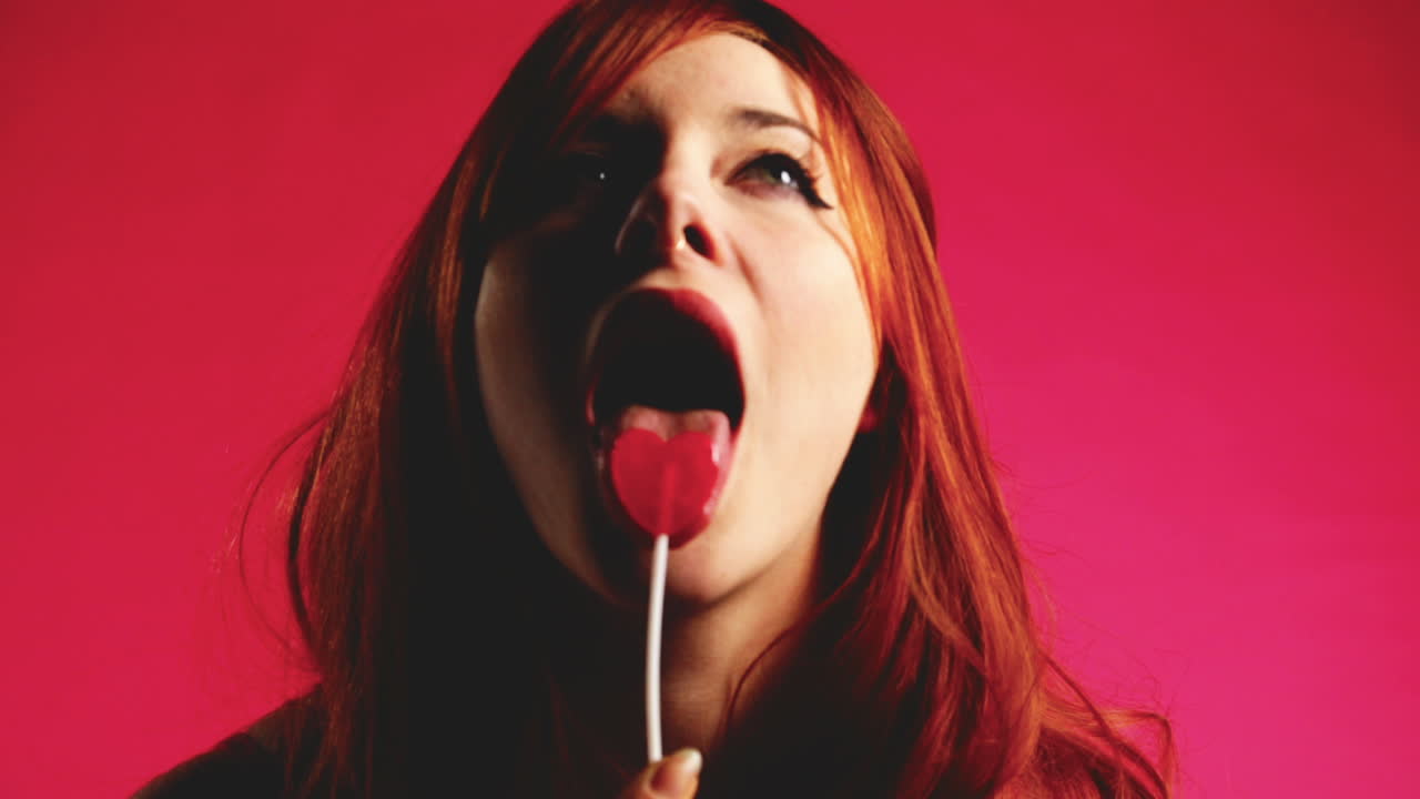 Close up of a green-eyed, red haired model, provocatively licks a heart shaped lollipop with her tongue in studio, on a pink backdrop