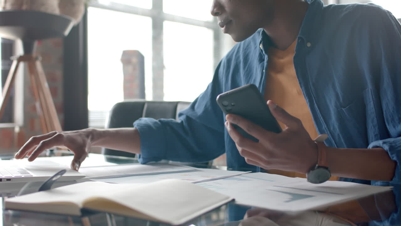 African american casual businessman sitting at desk using smartphone and laptop at home, slow motion
