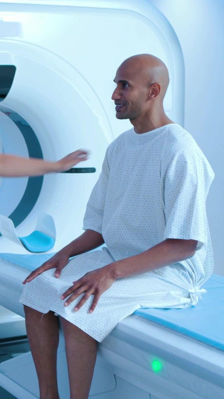 Video verticale: Paziente in camice riceve un tocco sulla spalla, si prepara per la scansione nella sala d'esame vicino al gantry.