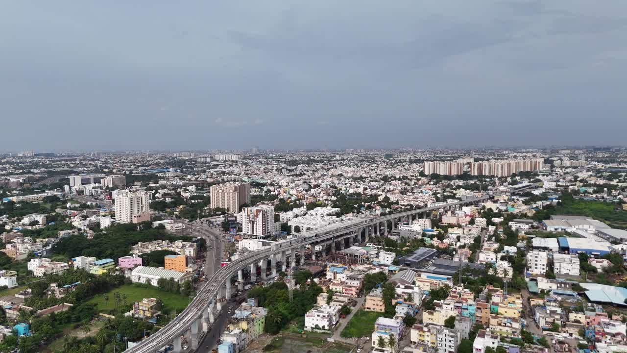 Wide aerial of Chennai urban sprawl. The elevated railway bisects a dense area of colorful low-rise Kattupakkam residences, contrasting with new, distant high-rise development under a dark sky