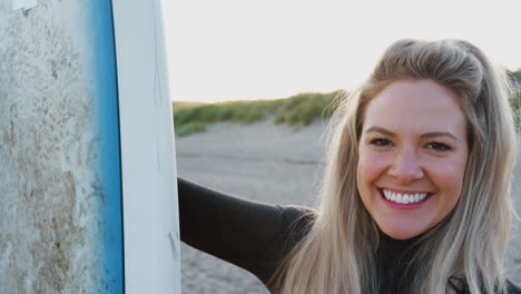 Portrait-Of-Woman-Wearing-Wetsuit-Holding-Surfboard-Enjoying-Surfing-Vacation-On-Beach-As-Sun-Sets