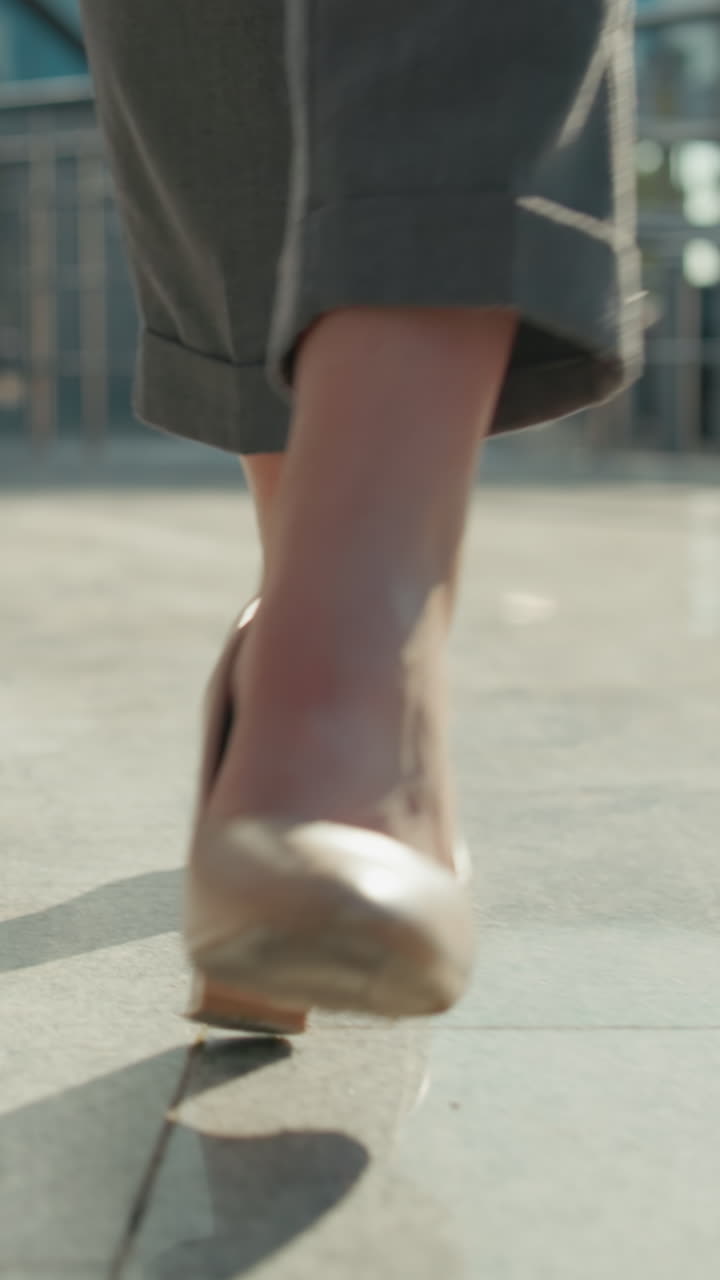 Close up leg view of confident lady walking gracefully in nude high heels on sunlit walkway with modern building, iron rail, parked cars in blurred background