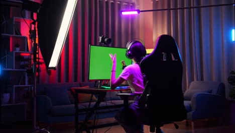 vista trasera de la chica asiática que transmite usando una computadora personal de pantalla verde, transmisión en vivo agitando la mano y hablando en el micrófono. escritorio iluminado por luz de tira led rgb
