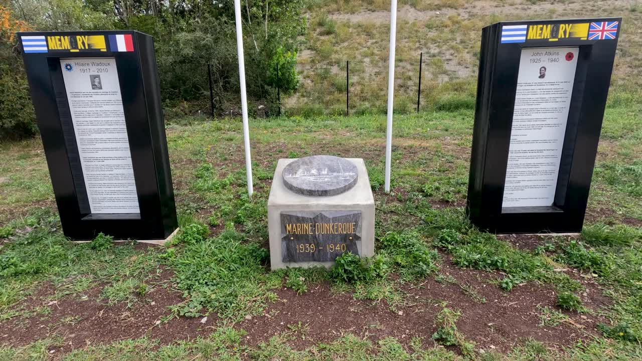 Two flags wave beside plaques and stone marker at outdoor war memorial in Dunkirk, France