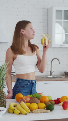 Girl eating banana indoors. Young woman on the modern kitchen background. Healthy fruit and vegetables on the table. Attractive female vegetarian is eating fruit. Vertical video