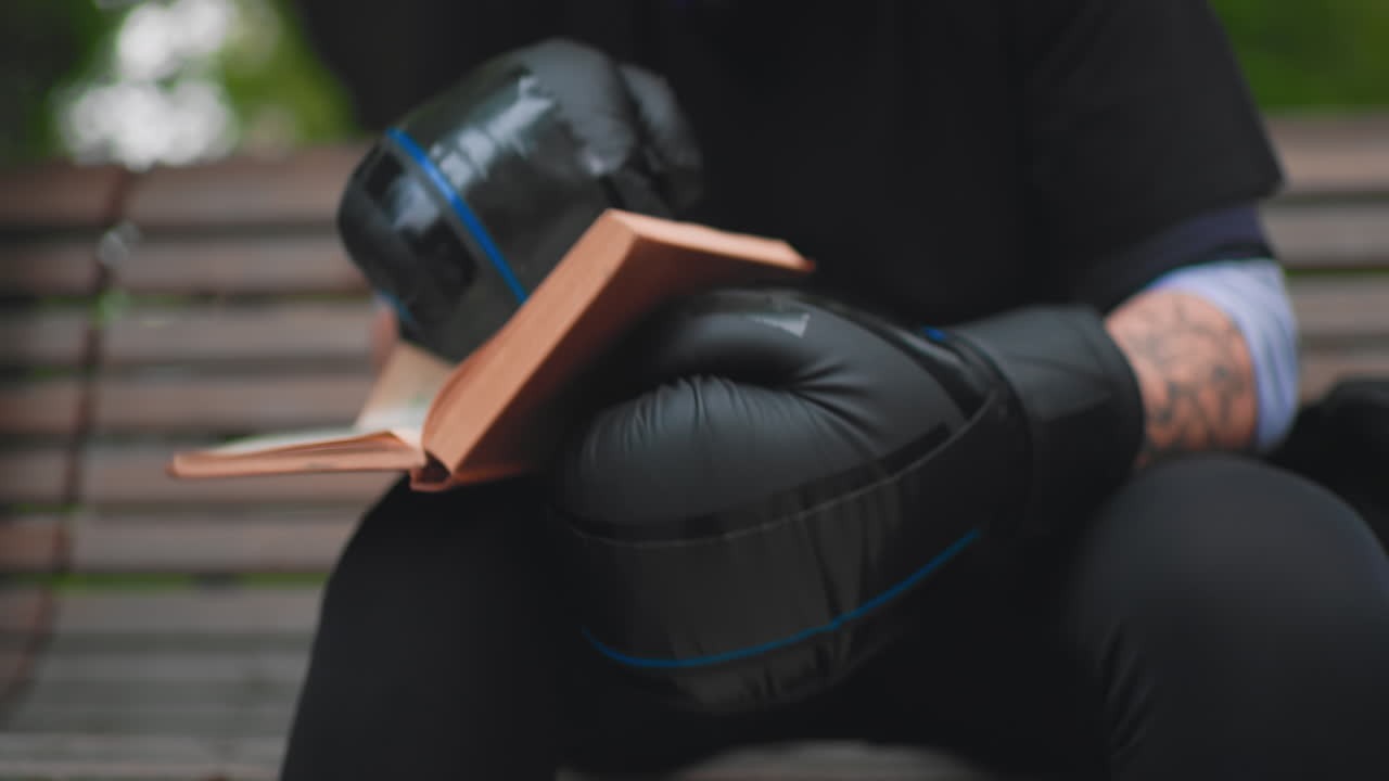 Persona assorta nella lettura, persona seduta su una panchina che si gode un momento di lettura tranquilla, lettore seduto su una panchina del parco perso nella contemplazione, uomo con braccia tatuate che legge con calma in un ambiente urbano