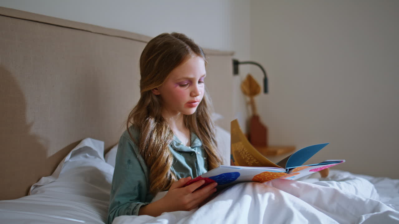 Bambina truccata che legge un libro e si rilassa sotto una coperta bianca, primo piano. Ragazzina a riposo